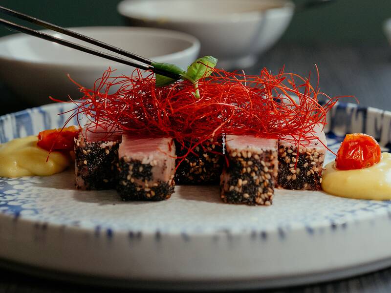 Ein kunstvoll angerichteter Teller mit Sushi-Würfeln, dekoriert mit dünnen roten Fäden und begleitet von einer gelben Sauce, wird elegant mit einer grünen Garnitur versehen.
