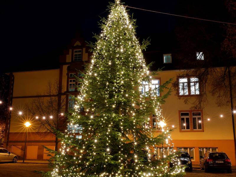 Ein großer, mit vielen Lichtern geschmückter Weihnachtsbaum steht nachts vor einem beleuchteten Gebäude, umgeben von Lichterketten.