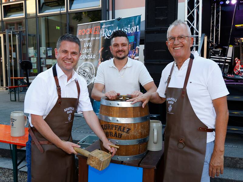 Drei Männer in Schürzen stehen vor einem Bierfass und lächeln in die Kamera, während sie scheinbar eine Bierveranstaltung veranstalten.
