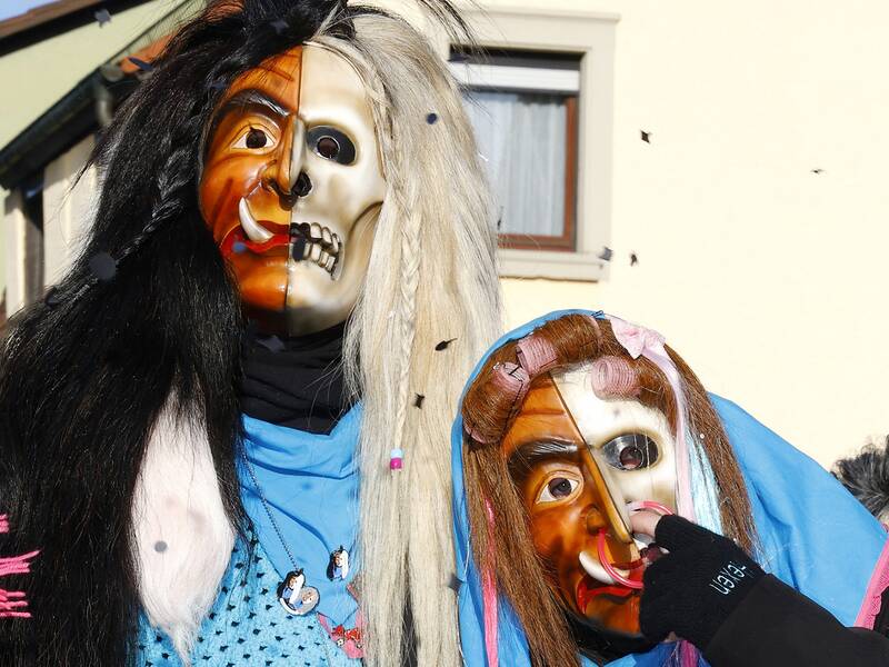 Zwei verkleidete Personen tragen bunte Masken, wobei eine Maske halb menschlich und halb skelettartig ist, während im Hintergrund ein Haus zu sehen ist.