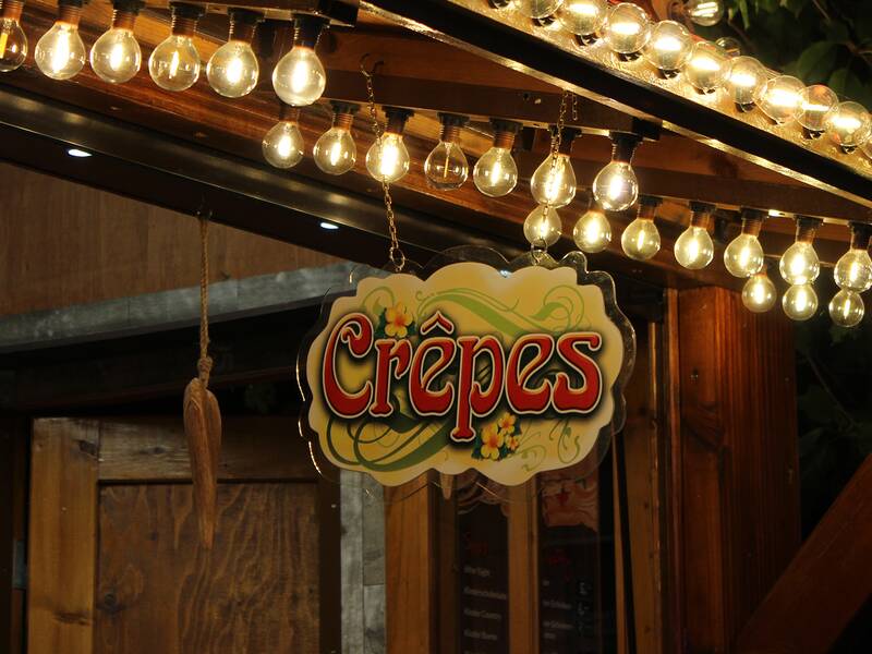 Ein Schild mit der Aufschrift "Crêpes" hängt unter einer Reihe von leuchtenden Glühbirnen an einem Holzstand.