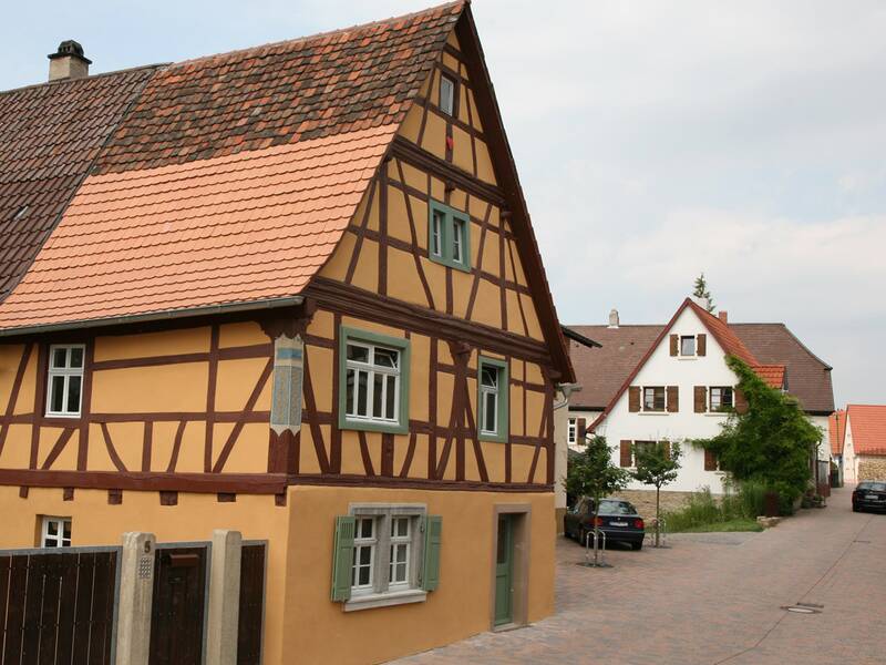 Ein Fachwerkhaus mit gelben Wänden und roten Ziegeldach steht in einer ruhigen, gepflasterten Straße neben weiteren Wohnhäusern.