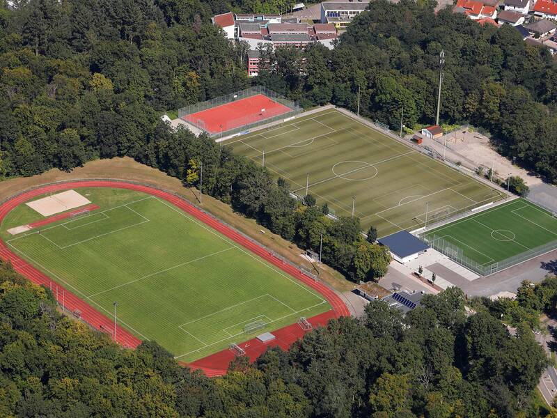 Das Bild zeigt eine Luftaufnahme eines Sportgeländes mit mehreren Sportplätzen, umgeben von Bäumen.