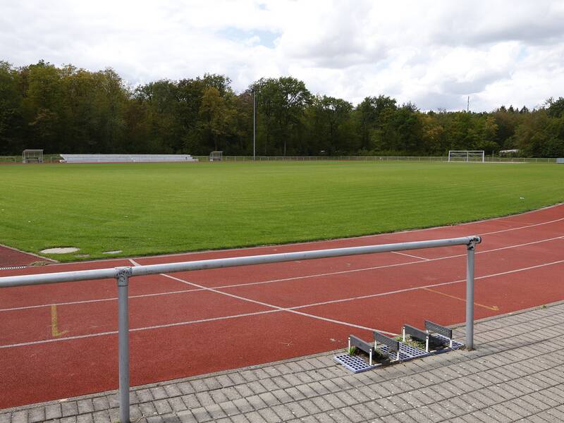 Das Bild zeigt ein leeres Sportstadion mit einer Laufbahn aus rotem Tartan, einem Fußballfeld in der Mitte und Bäumen im Hintergrund.