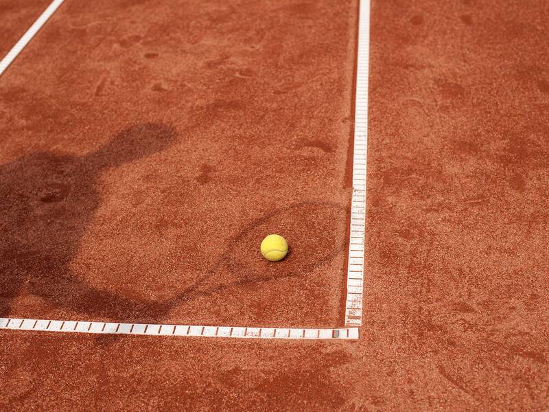 Ein Tennisball liegt auf einem Sandplatz in der Nähe der Linien, und der Schatten einer Person ist zu sehen.