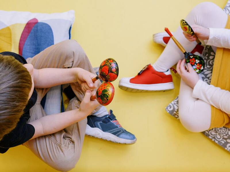 Zwei Kinder sitzen auf einem gelben Boden und halten bunte, verzierte Rasseln in den Händen.