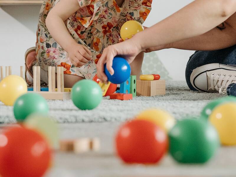Eine Person und ein Kind spielen auf einem Teppich mit bunten Bällen und Bauklötzen.