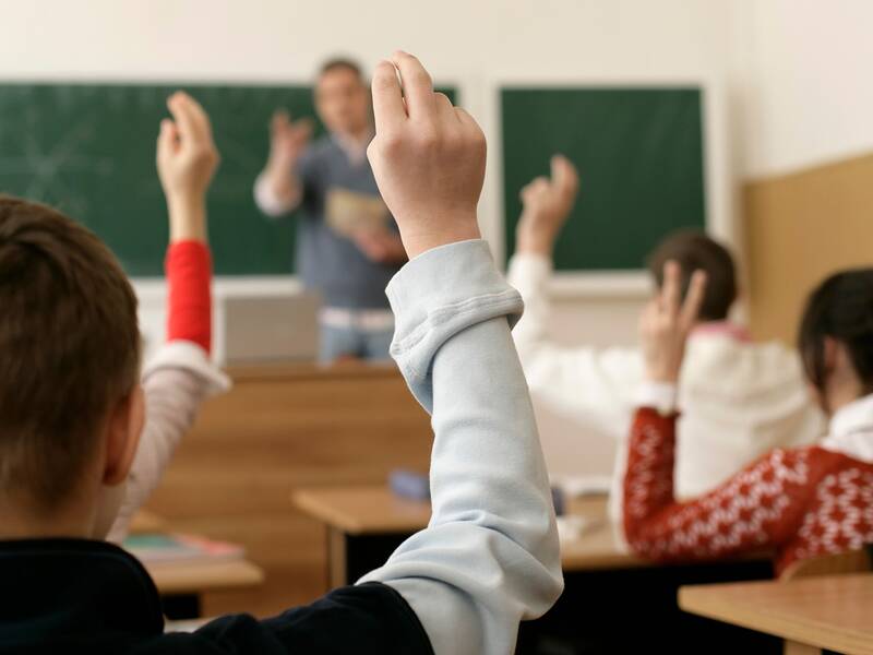 Mehrere Schüler in einem Klassenzimmer heben die Hand, während ein Lehrer vor einer Tafel steht.