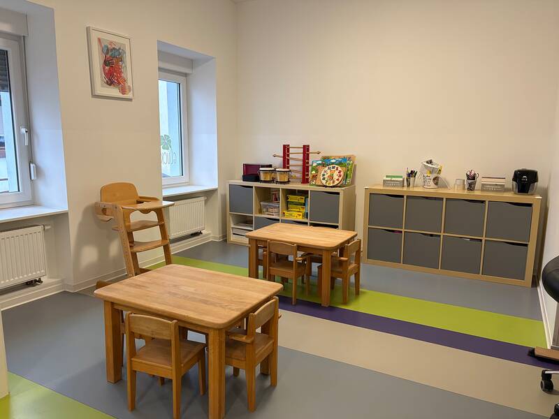 Ein heller Raum mit zwei kleinen Holztischen und Kinderstühlen, einem Regal voller Spielzeuge und zwei Fenstern mit einem Kunstwerk an der Wand.