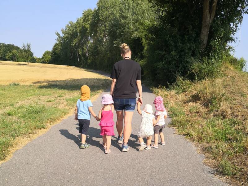 Eine erwachsene Person geht mit vier kleinen Kindern, die Hüte tragen, einen schmalen Weg entlang, der von Bäumen und Feldern gesäumt ist.