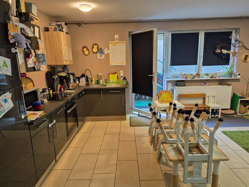 Ein heller Raum mit einer Küchenzeile auf der linken Seite, einem Tisch mit darauf gestapelten Kinderstühlen in der Mitte und einem Fenster mit schwarzen Jalousien im Hintergrund.