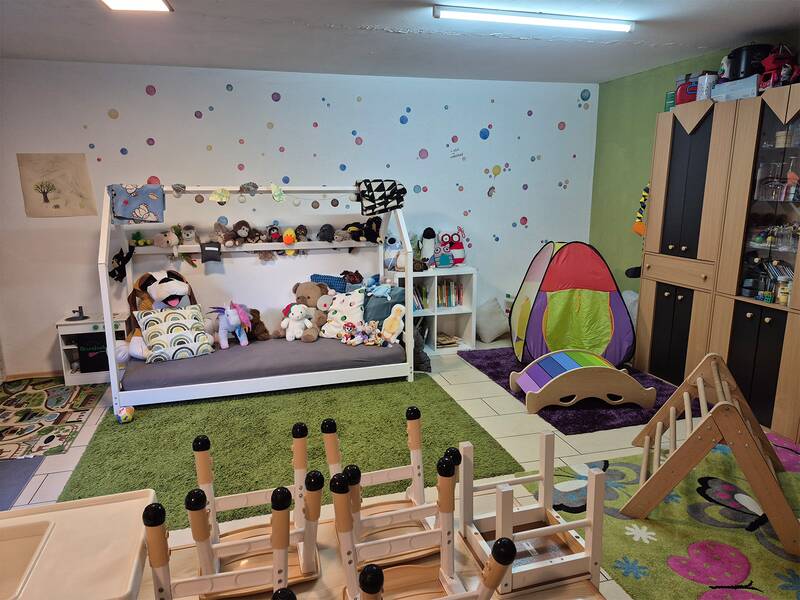 Der Raum ist ein bunt dekoriertes Kinderzimmer mit vielen Stofftieren auf einem Sofa, einem kleinen Zelt, Spielgeräten und einem Teppich auf dem Boden.
