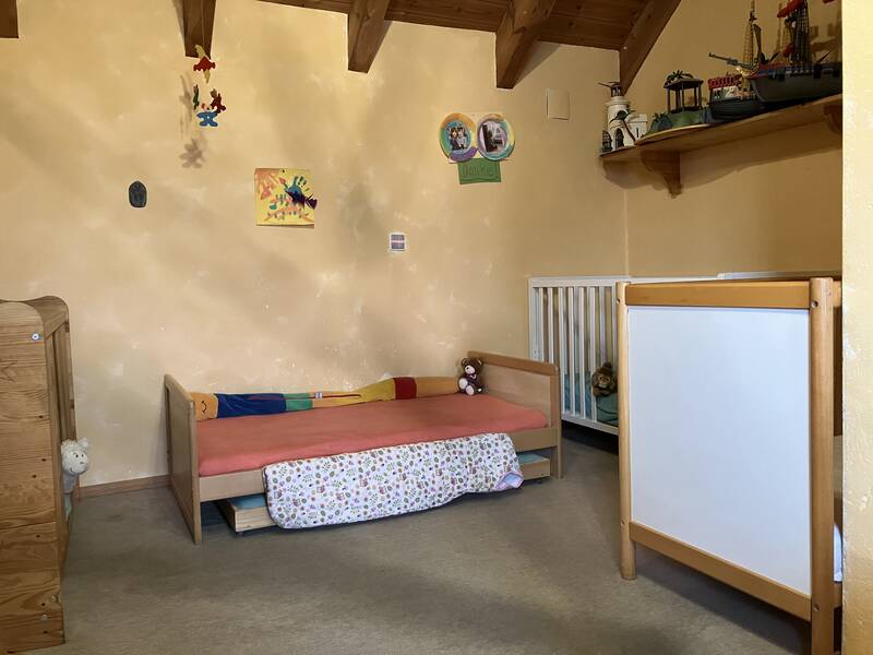 Ein Kinderzimmer mit einem Holzbett, einem Gitterbett, farbigen Mobiles und einem Spielzeugschiff auf einem Wandregal.