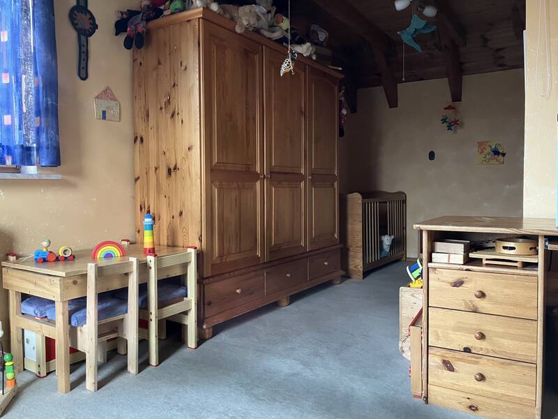 Ein Kinderzimmer mit Holzmöbeln, darunter ein Tisch mit zwei Stühlen, ein großer Schrank, ein Gitterbett und diverse Spielzeuge.