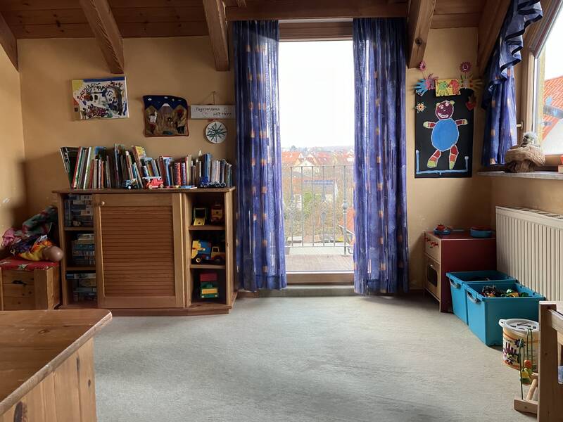Ein Kinderzimmer mit einem Holzschrank voller Bücher und Spielzeug, einem großen Fenster mit blauen Vorhängen und farbenfrohen Kinderzeichnungen an den Wänden.