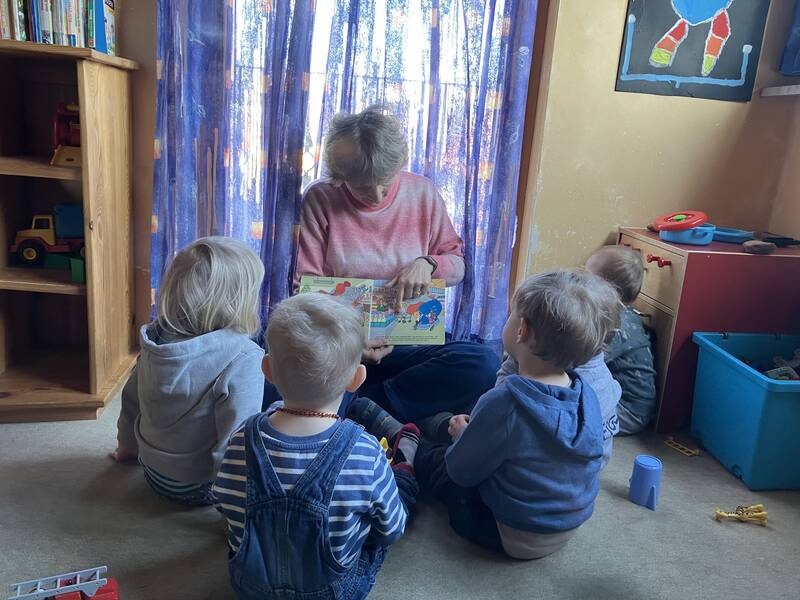 Eine Person liest mehreren kleinen Kindern in einem Zimmer mit Spielzeugen und bunten Vorhängen ein Bilderbuch vor.