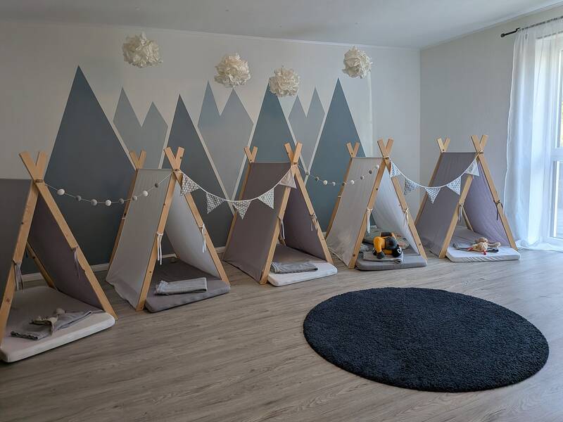 Eine gemütliche Ecke mit fünf kleinen Indoor-Zelten, die Holzböden, kleine Matratzen und eine dekorative Lichterkette sowie ein rundes, dunkles Teppichstück zeigen, wird von einer Wand mit Bergmotiven im Hintergrund ergänzt.