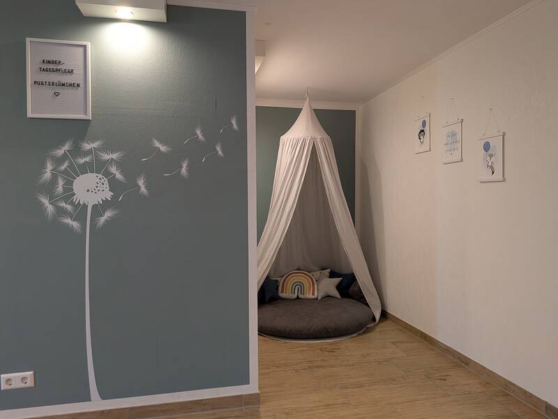 Ein gemütlicher Raum mit einem mintgrünen Wandbereich, auf dem eine Pusteblume abgebildet ist, und einem kleinen, mit Kissen ausgestatteten Spielzelt in der Ecke.