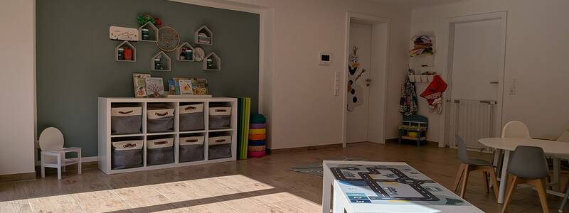 Ein heller Raum mit Holzboden, in dem sich ein weißer Tisch mit Spielmatte, Kinderstühle, ein Regal mit Körben und Wanddekorationen befinden.