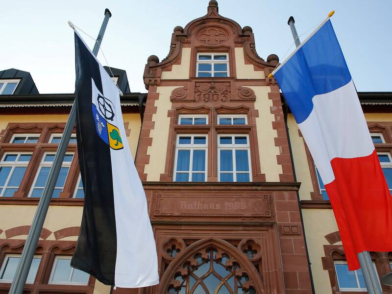 Vor einem historischen Gebäude mit der Inschrift "Rathaus 1903" wehen zwei Fahnen, die eine in Blau-Weiß-Rot und die andere in Schwarz-Weiß mit einem Wappen.