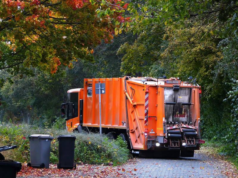 Ein orangefarbenes Müllfahrzeug fährt eine von Bäumen gesäumte Straße entlang, während drei Mülltonnen am Straßenrand stehen.