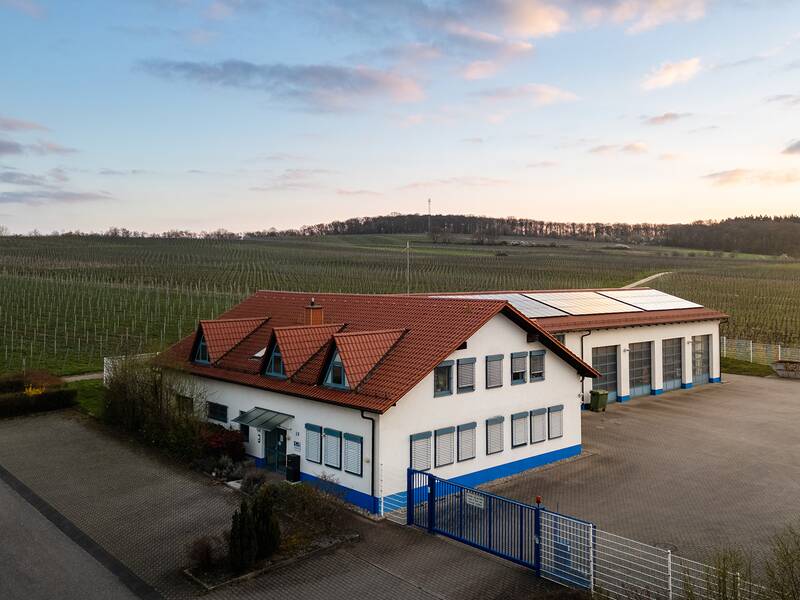 Eine weiß-blaue Lagerhalle mit roten Dachziegeln steht neben einer großen, gepflasterten Fläche und ist von weitläufigen, grünen Feldern umgeben.