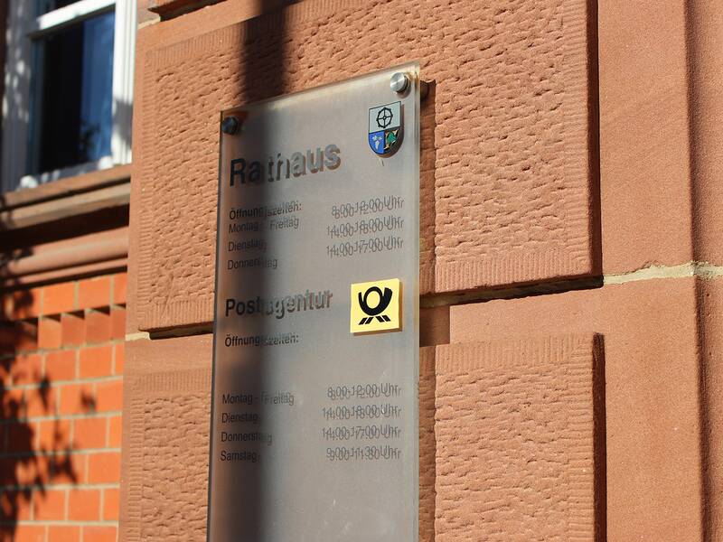 Ein Schild an einer Backsteinmauer zeigt die Öffnungszeiten eines Rathauses und einer Postagentur.