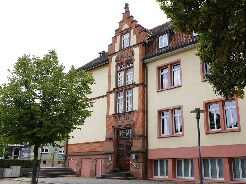 Ein historisch anmutendes Schulgebäude mit rotbraunen Ziegeln und gelben Wänden, flankiert von Bäumen.