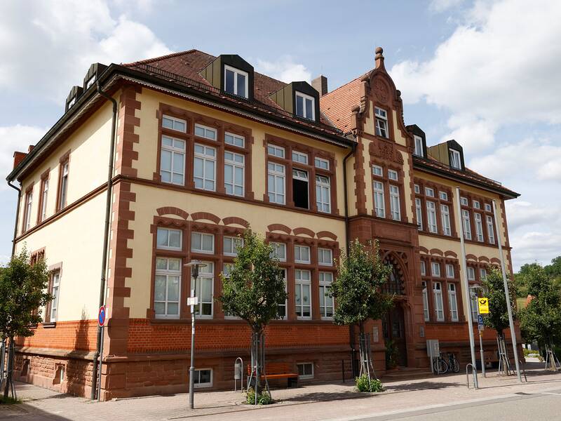 Das Bild zeigt ein historisches Gebäude mit rotbraunen Zierelementen und großen Fenstern, umgeben von kleinen Bäumen auf einem städtischen Gehweg.