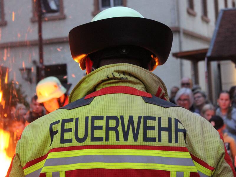 Ein Feuerwehrmann in Schutzkleidung steht vor einem lodernden Feuer, während im Hintergrund eine Menschenmenge zuschaut.