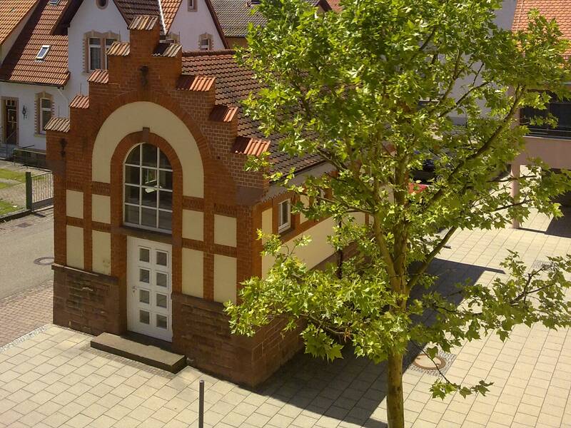 Ein kleines gemauertes Gebäude mit einer dekorativen Fassade steht in einer gepflasterten Umgebung, daneben ein grüner Baum, vor einem klaren blauen Himmel.