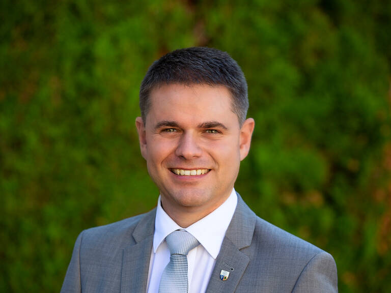 Ein lächelnder Mann in einem grauen Anzug steht vor einem verschwommenen grünen Hintergrund.