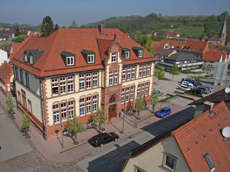 Das Bild zeigt ein historisches Gebäude mit rotem Dach inmitten einer Kleinstadt, umgeben von Straßen, Bäumen und parkenden Autos.