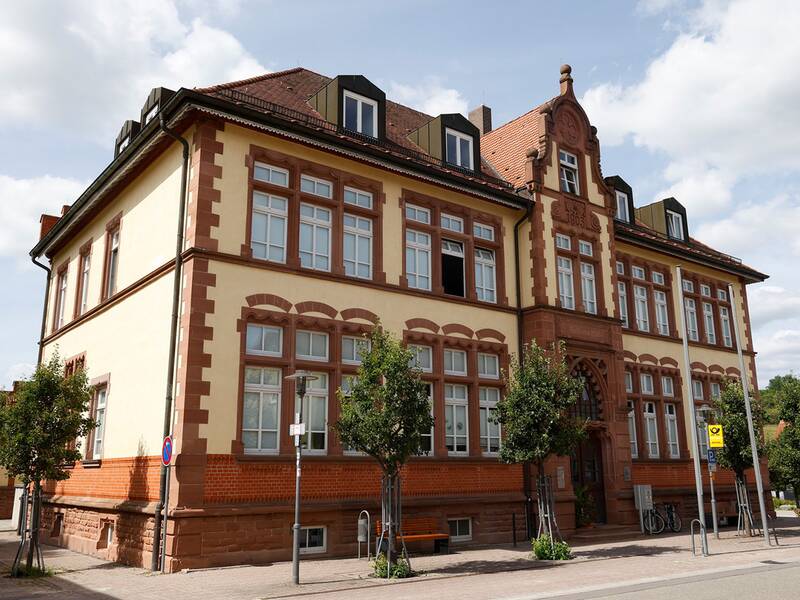 Ein historisches Gebäude mit roten Ziegelverzierungen und großen Fenstern steht an einer Straße, umgeben von Bäumen und einem Wartehäuschen.