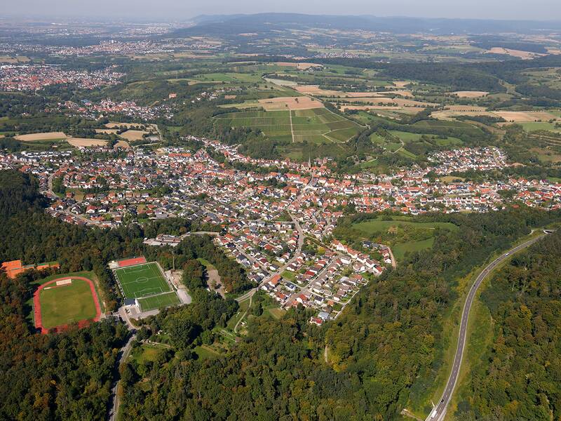 Luftaufnahme eines Dorfes mit vielen Häusern, umgeben von grünen Feldern, Wäldern und Sportplätzen.