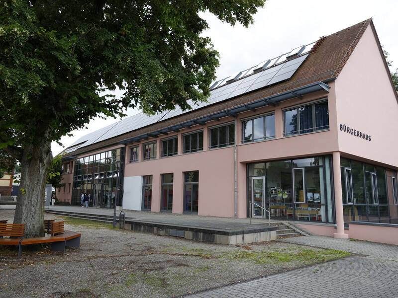 Ein modernes Gebäude mit der Aufschrift "Bürgerhaus" und Solarpanels auf dem Dach steht neben großen Bäumen und einer gepflasterten Fläche.