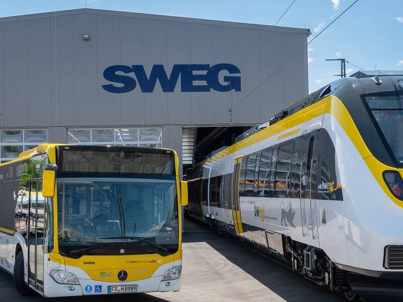 Ein gelber Bus und ein weiß-gelber Zug stehen vor einem Gebäude mit der Aufschrift "SWEG".