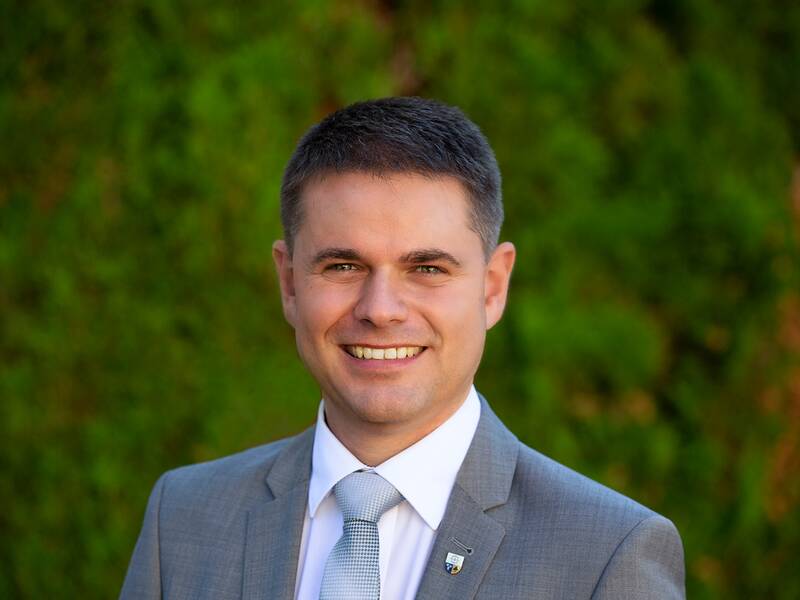 Ein lächelnder Mann in einem grauen Anzug mit Krawatte steht vor einem verschwommenen, grünen Hintergrund.